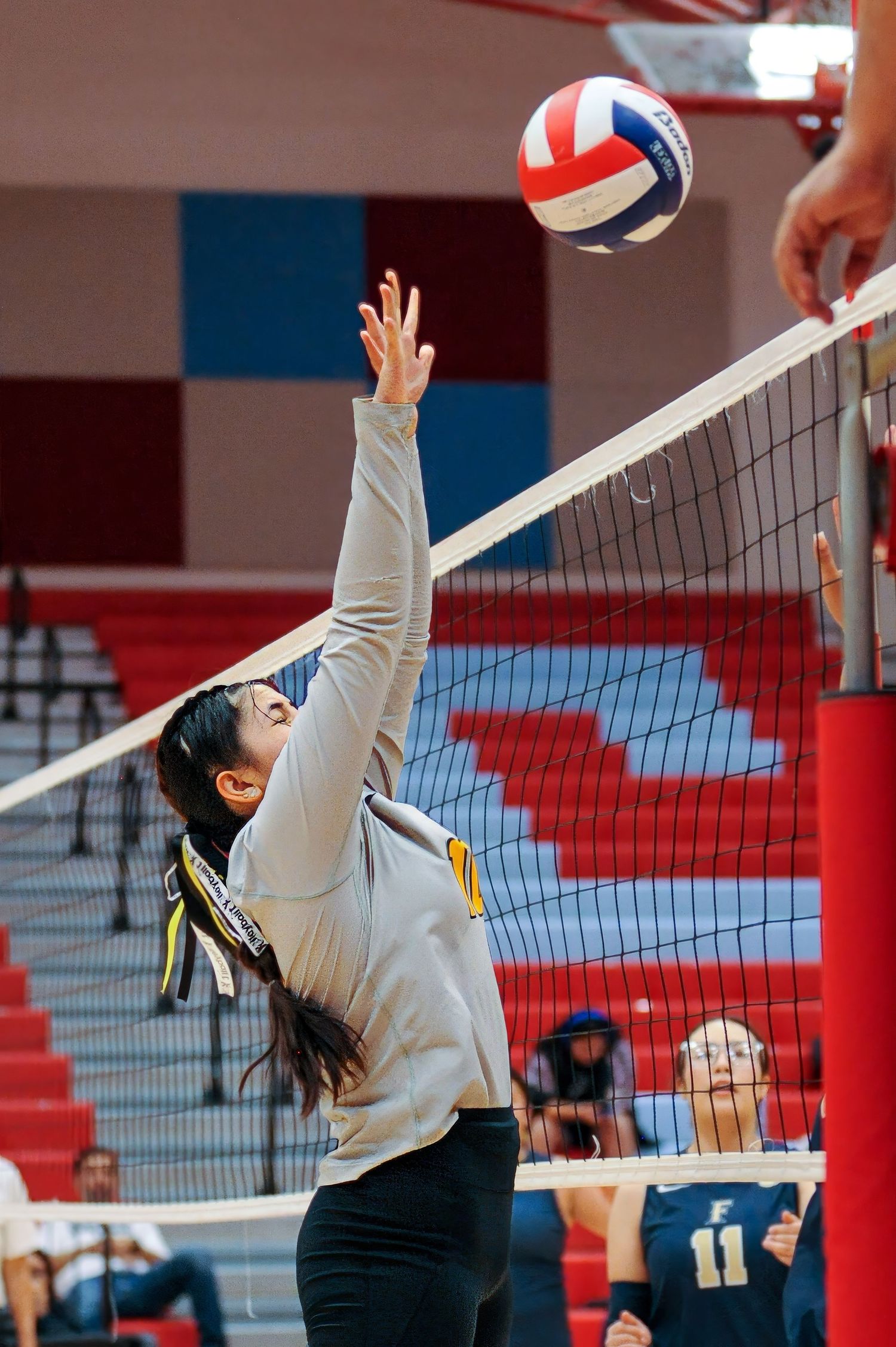 Female volleyball player jumping to spike ball over net during indoor match in red gymnasium.