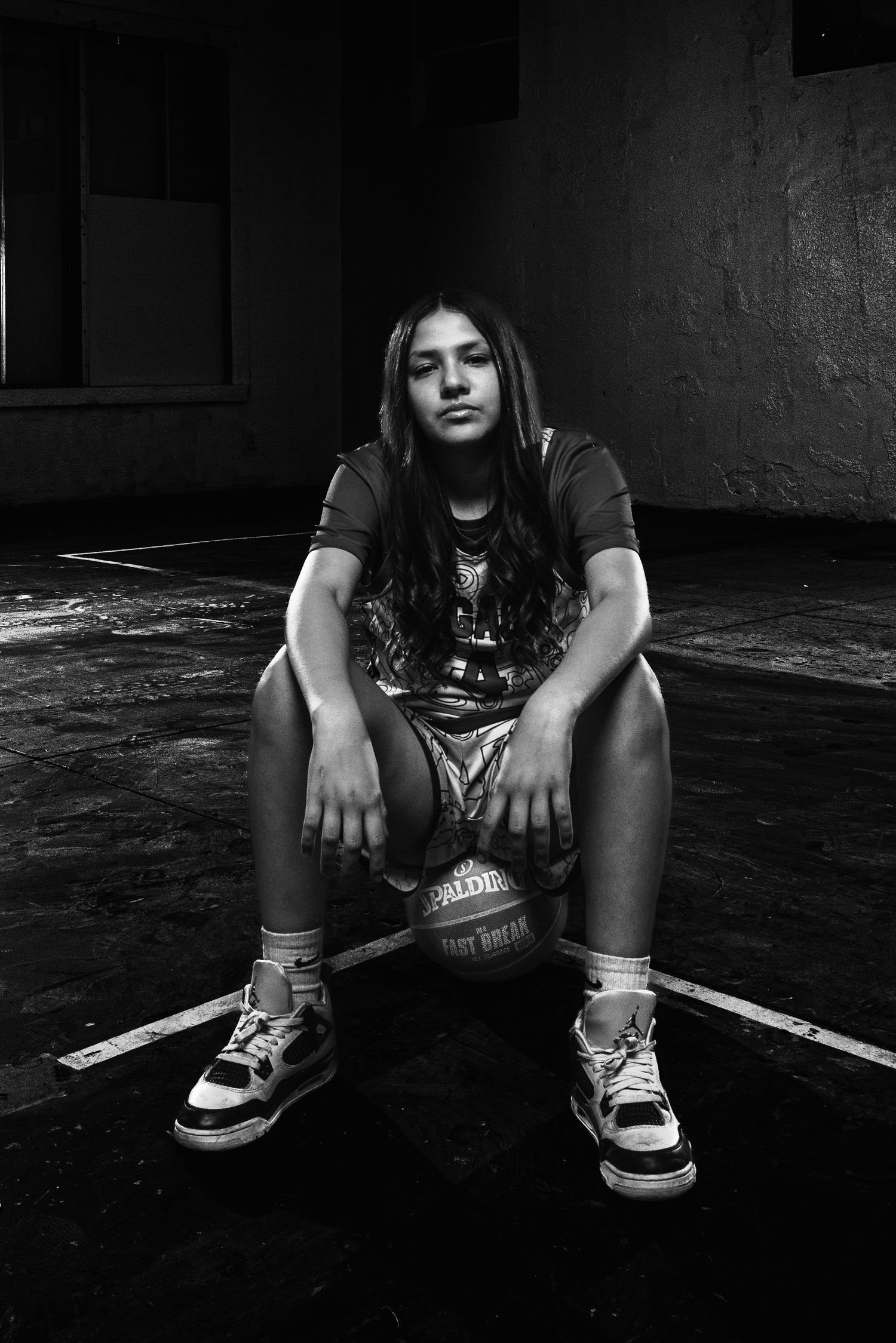 Black and white photo of a young girl sitting on a Spalding basketball on an indoor court.