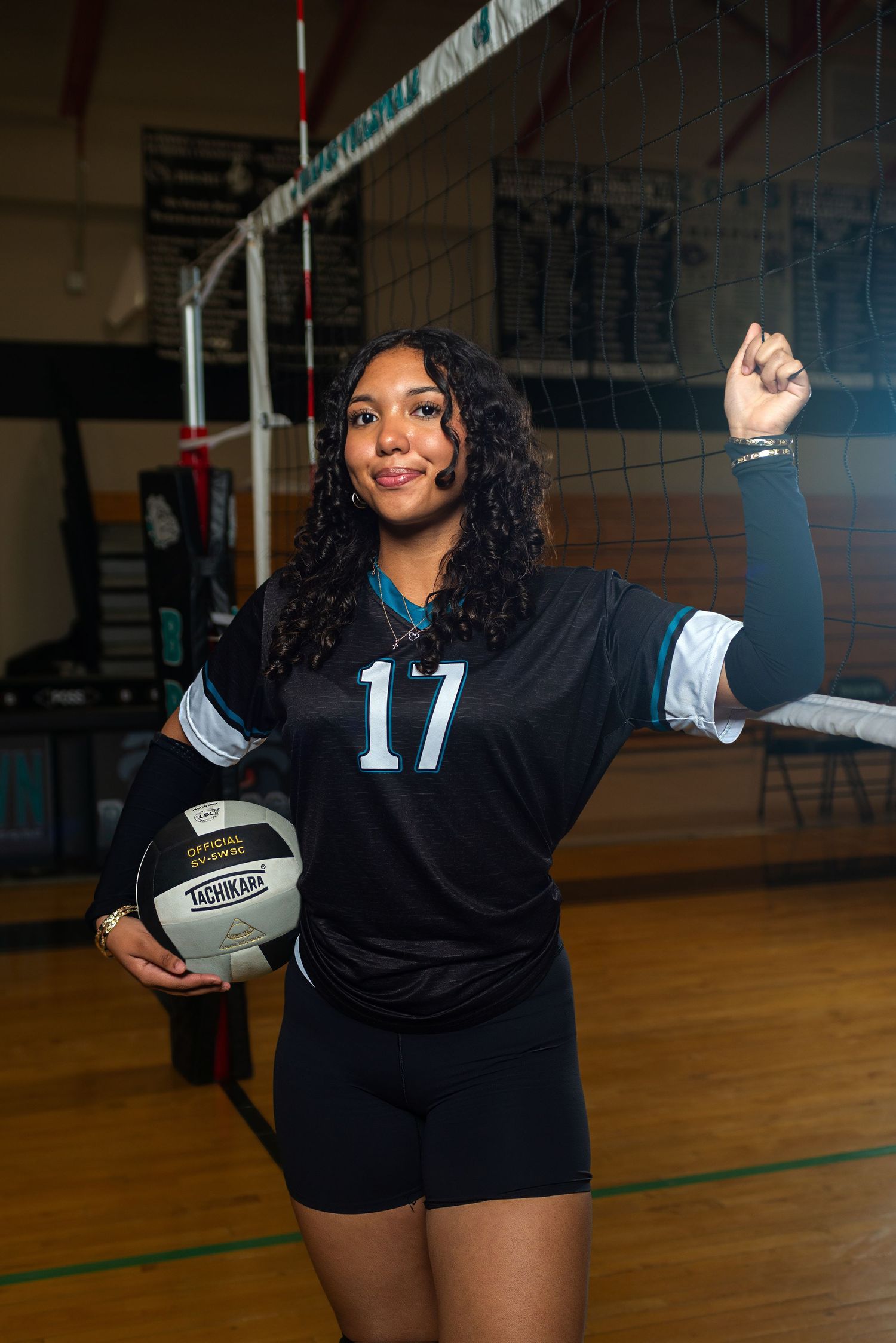 Female volleyball player #17 poses confidently in black jersey, holding ball with fist raised in gym.