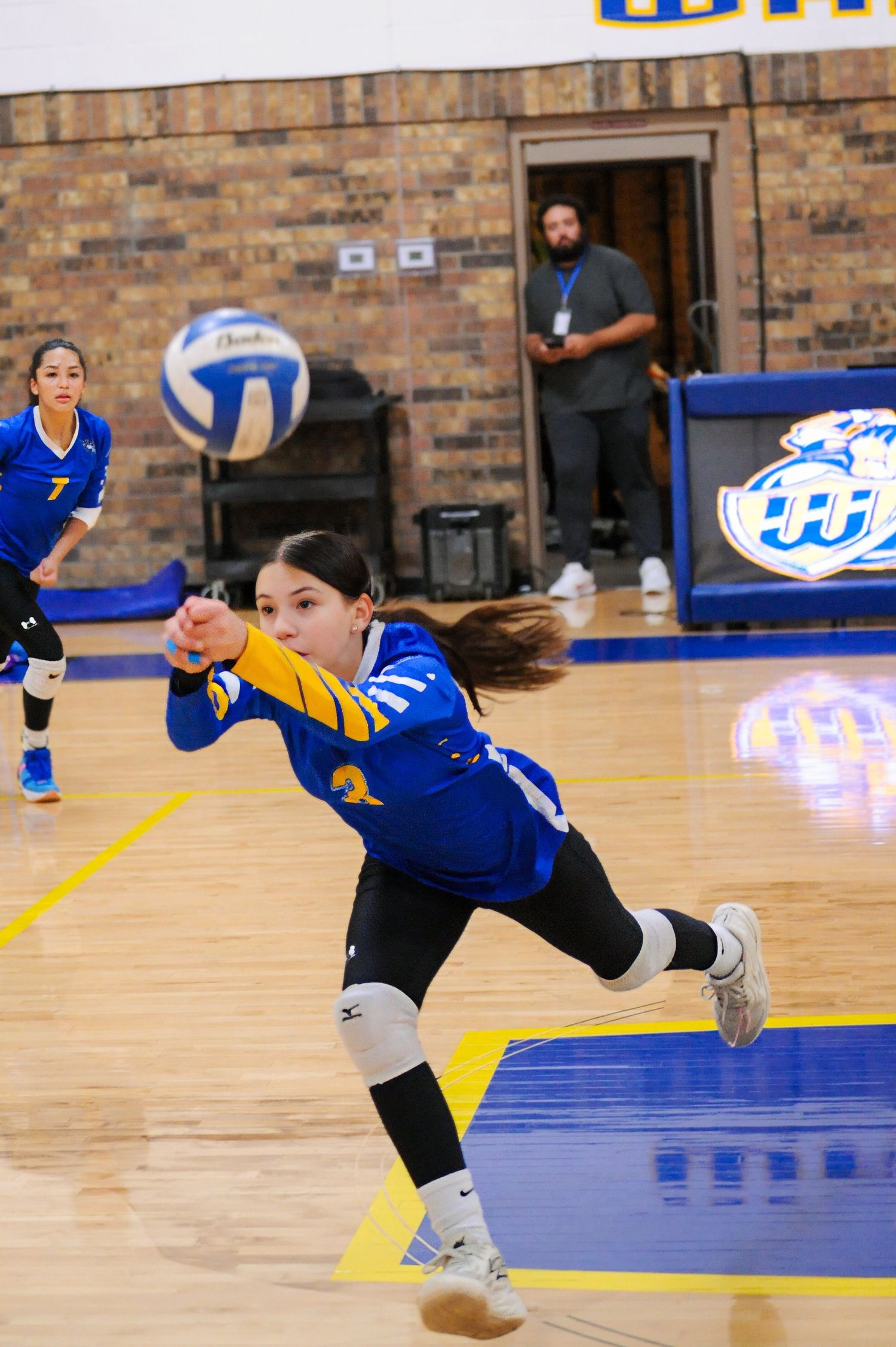 A volleyball player in a blue and gold uniform digs the ball during an indoor match with teammates visible behind her.