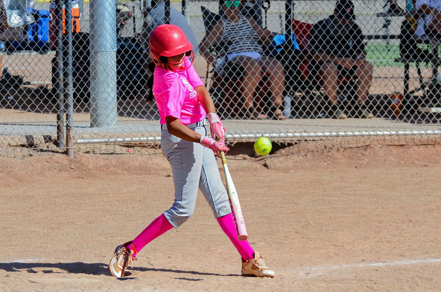 Young softball batter in pink uniform and red helmet swinging at a yellow pitch during an outdoor game.