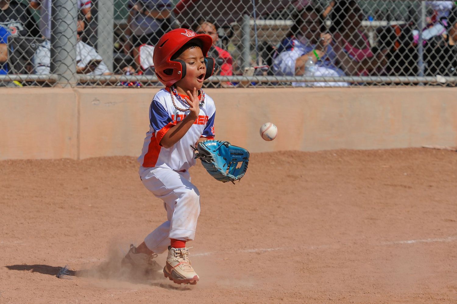 Young boy in red helmet catches baseball with blue glove during Little League game on dirt field.