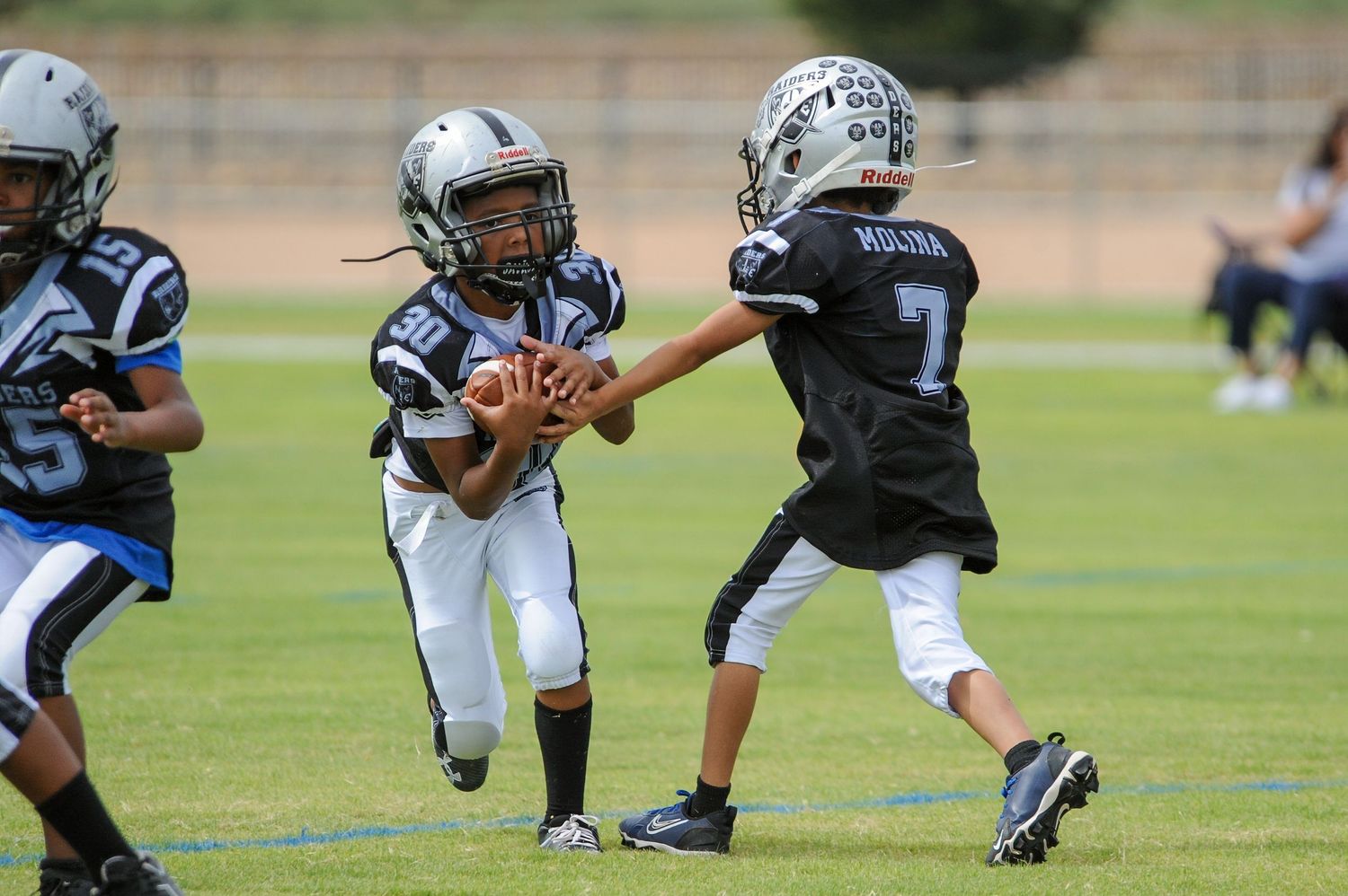 Two youth football players in black Raiders uniforms exchange a handoff during a youth league football game outdoors.