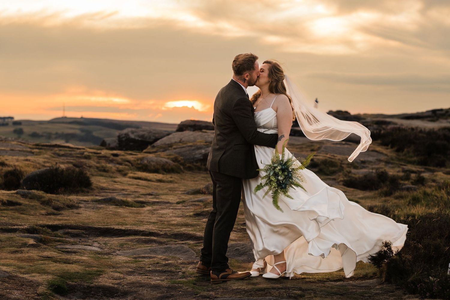 Norfolk wedding photographer. A couple shares an intimate moment at sunset on rocky terrain as the bride's white dress flows in the wind.
