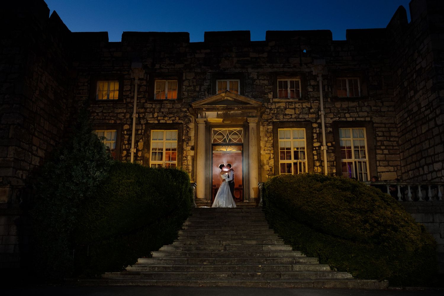 Dramatic wedding portrait of bride and groom in front of a castle with illuminated windows and steps - by Norfolk Wedding Photographer.