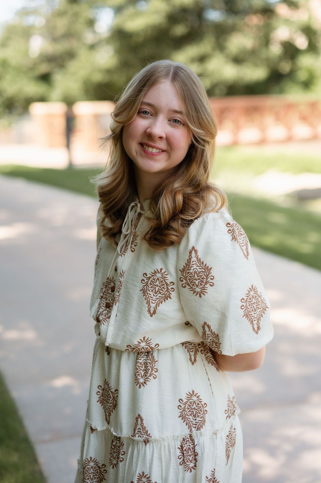 Smiling young woman in white paisley dress poses on a sunny garden pathway surrounded by trees.