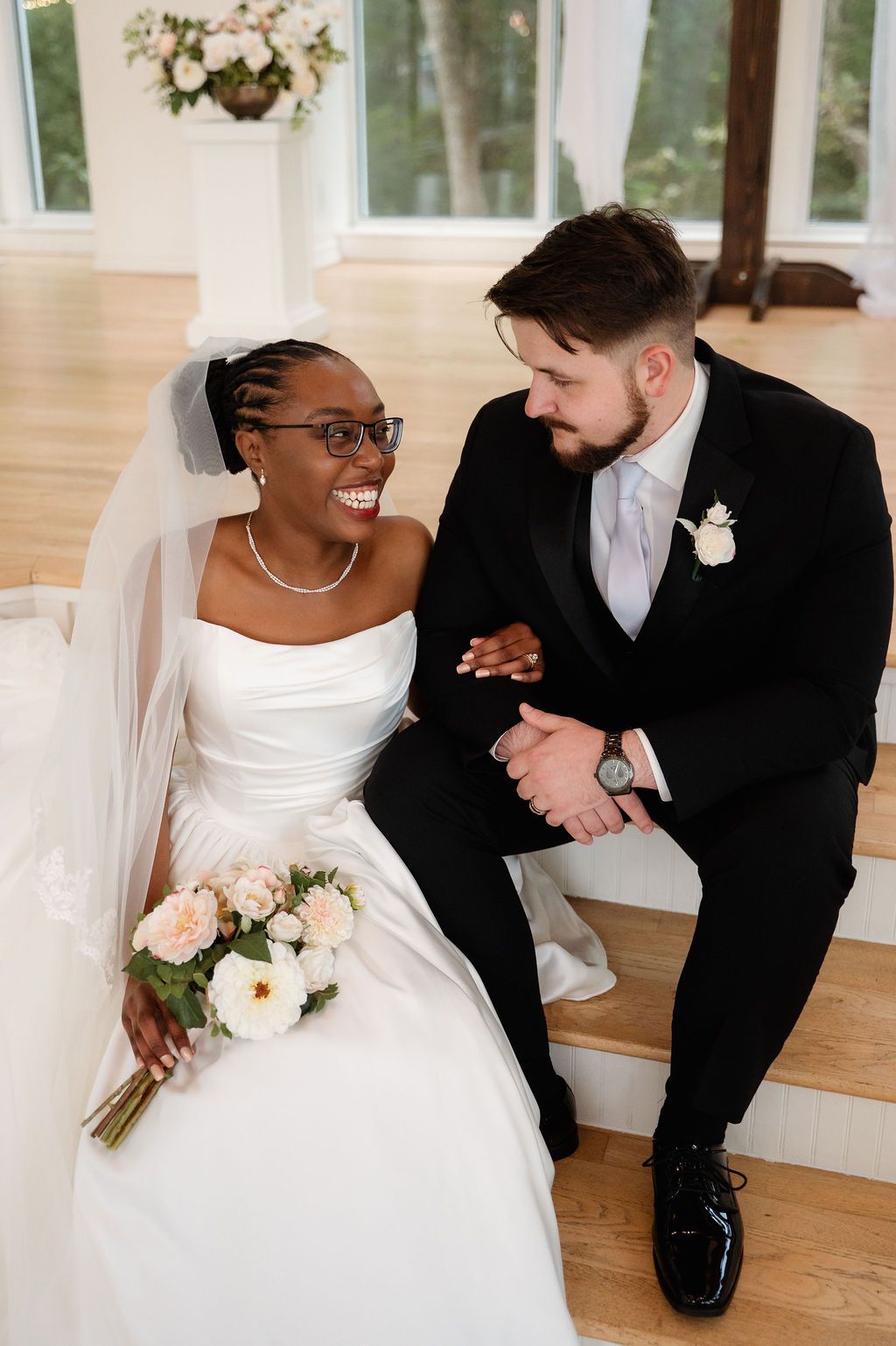 Interracial bride and groom sitting on stairs laughing together, bride holding white bouquet in wedding venue.