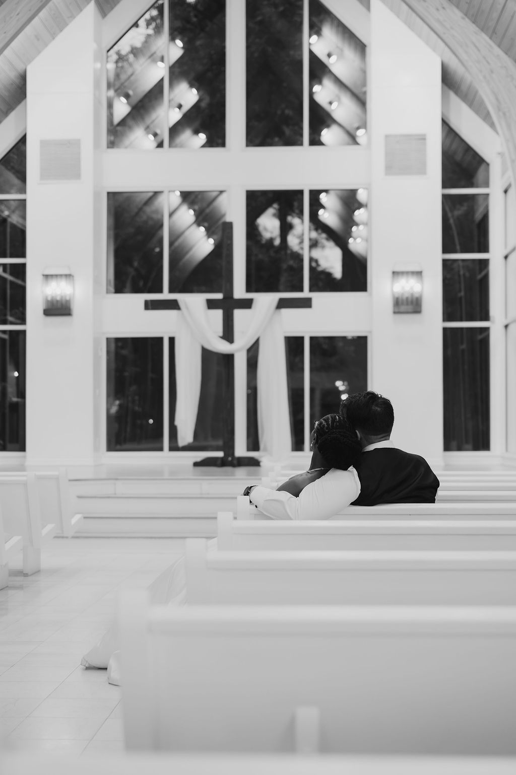 Black and white photo of a couple sitting together in a white chapel with a cross and large windows.