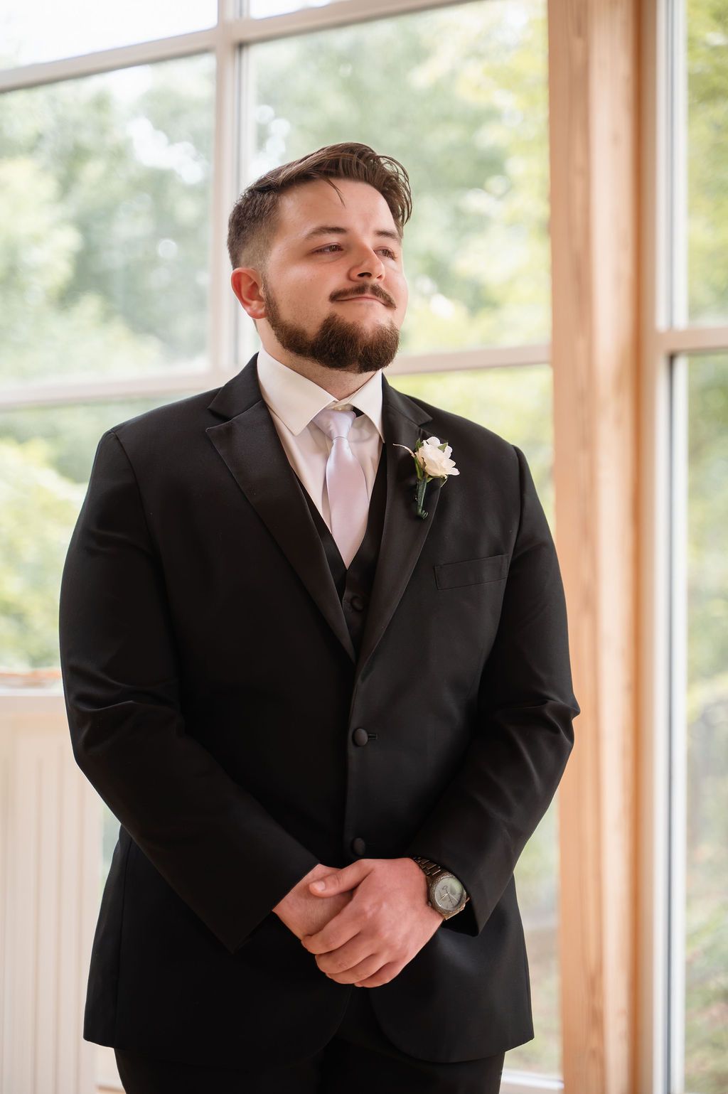 Groom in black tuxedo with white boutonniere and pink tie stands clasping hands before large windows.