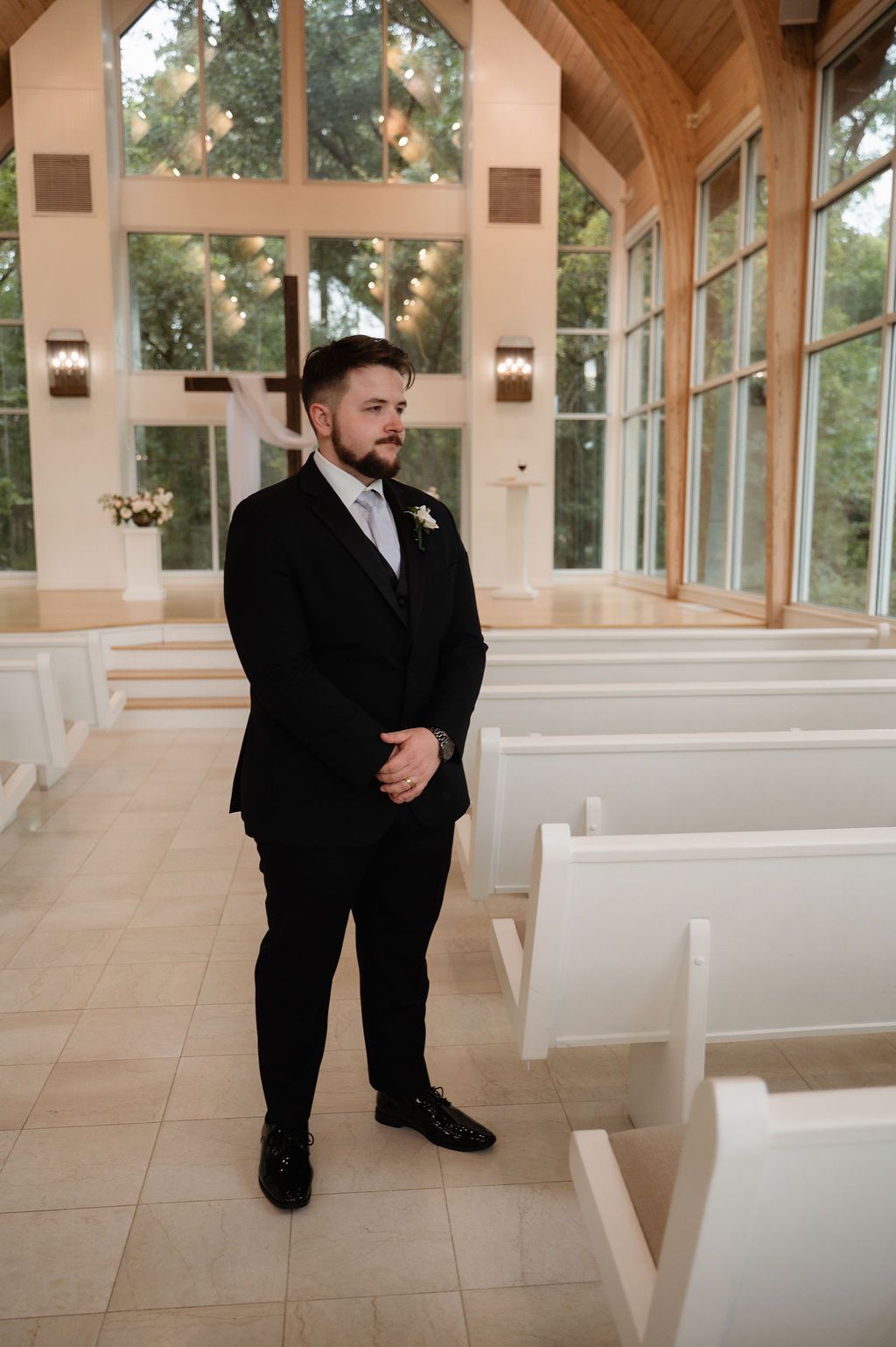 Groom in black suit standing at chapel altar with white pews and large windows in bright wedding venue.