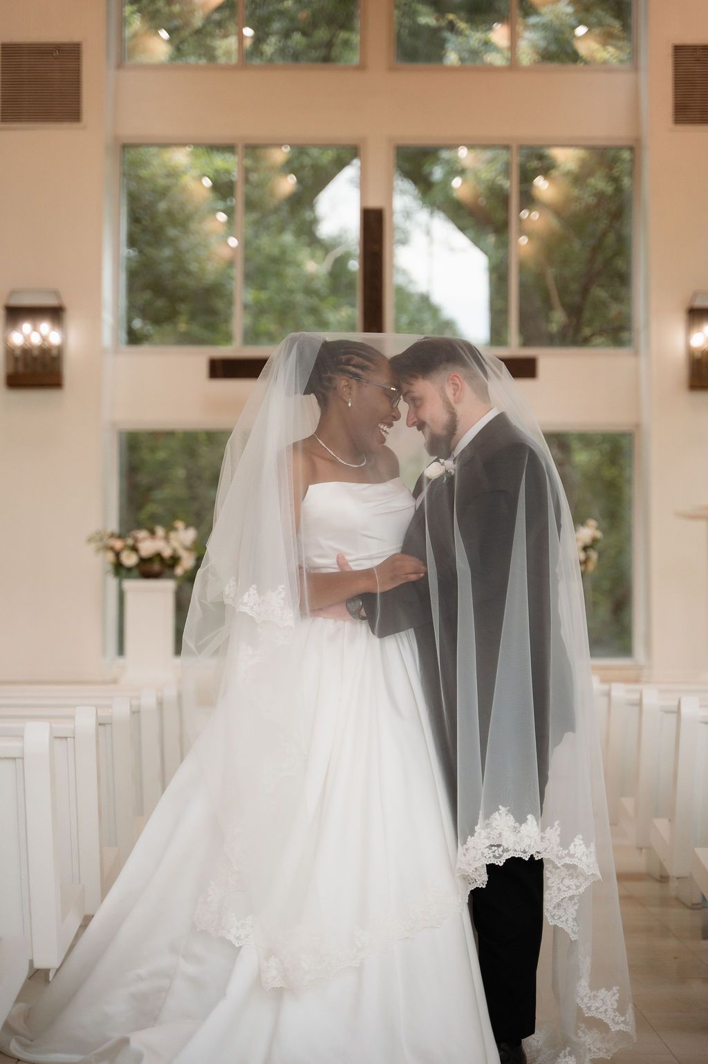 Bride and groom sharing intimate moment under lace veil in white chapel with cross and large windows.