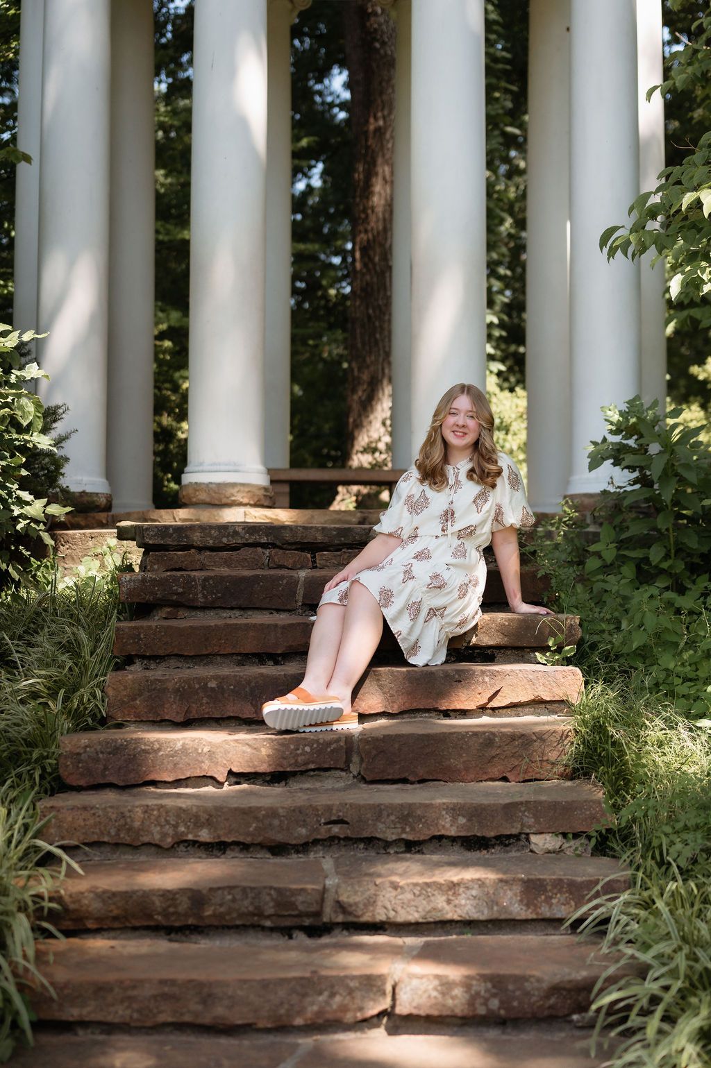 Smiling teen girl in floral dress sits on stone steps near white columns for senior photos.