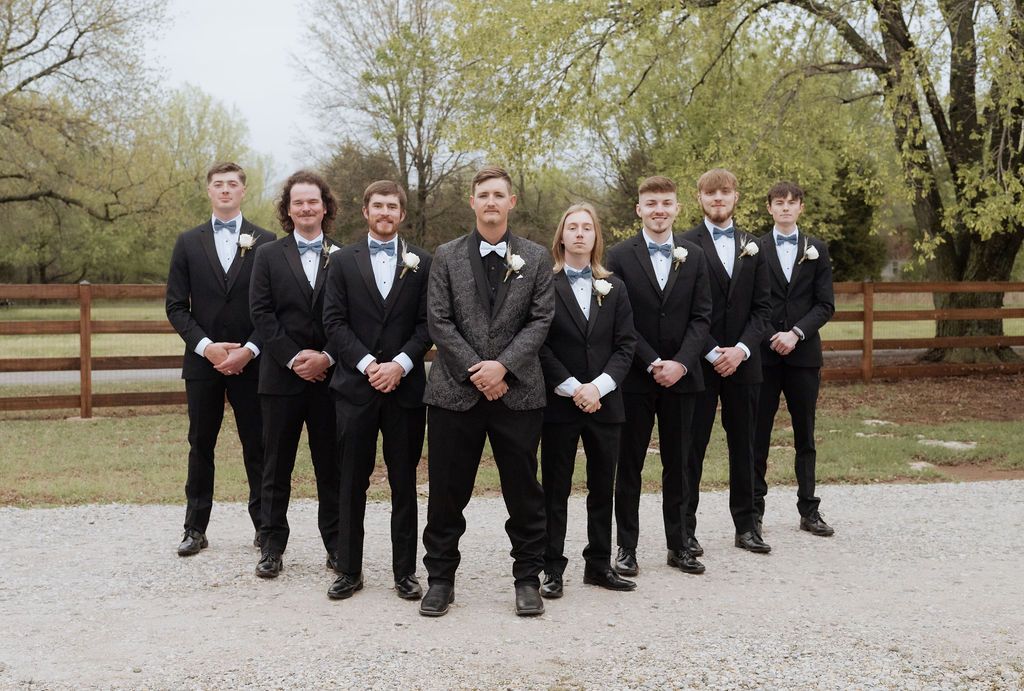 Group of seven young men in black tuxedos with bow ties posing outdoors at a Tulsa area Wedding
