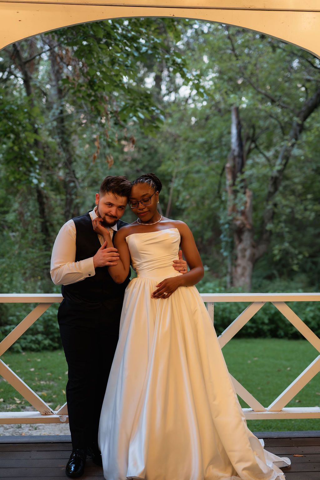 Interracial couple poses elegantly outdoors in wedding attire, bride in white gown, groom in black vest, under arched gazebo.