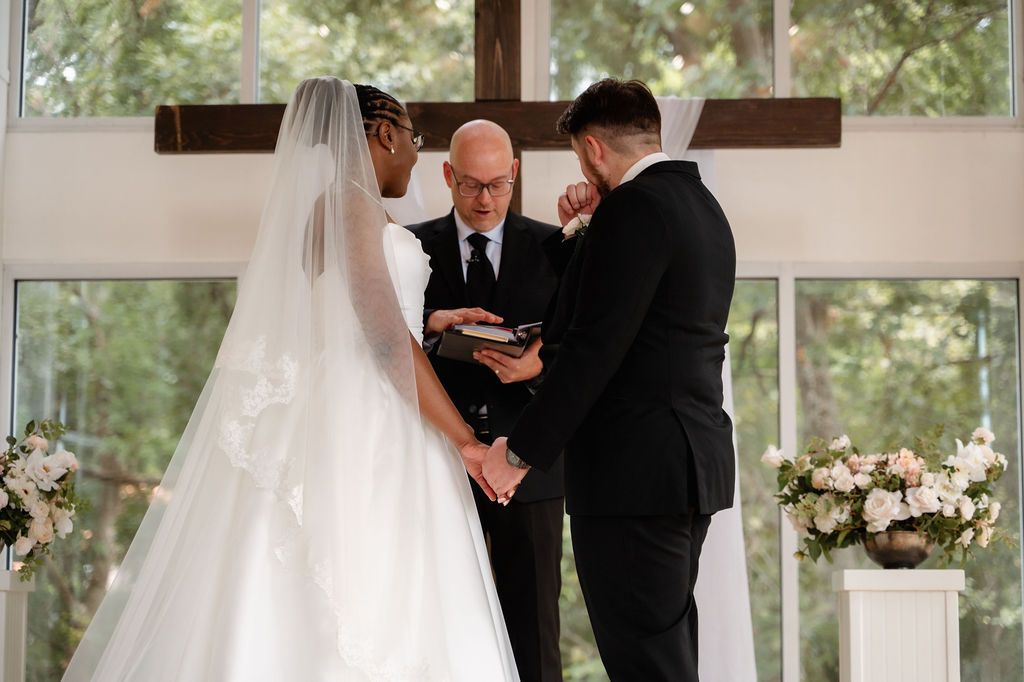 Bride and groom exchange vows at elegant chapel altar with large cross, officiant holding ceremony book, floral arrangements nearby.