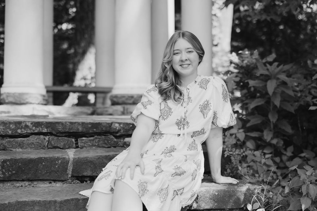 Black and white senior portrait of smiling girl in floral dress seated on stone steps by columns.