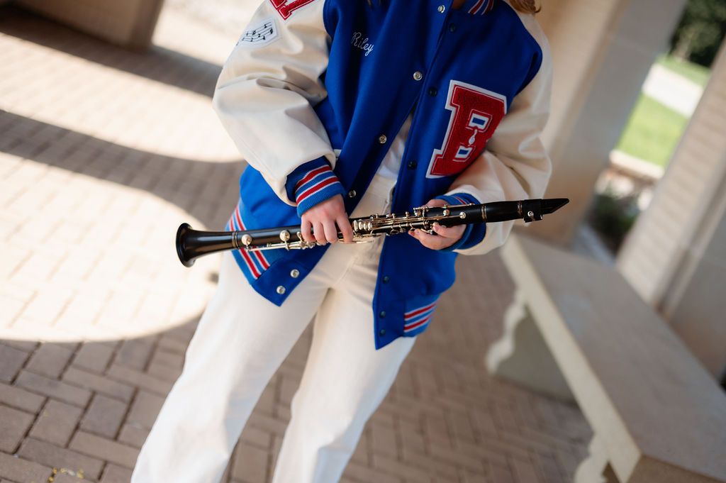 Close-up of person in varsity jacket holding a black clarinet on a brick patio.