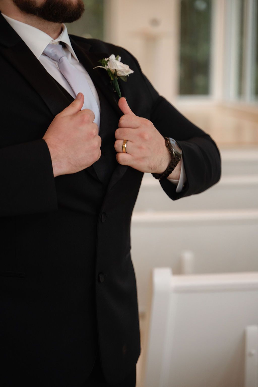 Groom in black suit adjusting lapel with white boutonniere, gold wedding ring, and watch, standing in a chapel.