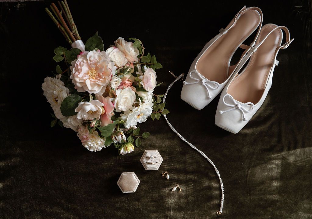 Bridal flat shoes with white bows beside a pink peony bouquet and hexagonal ring box on dark surface.