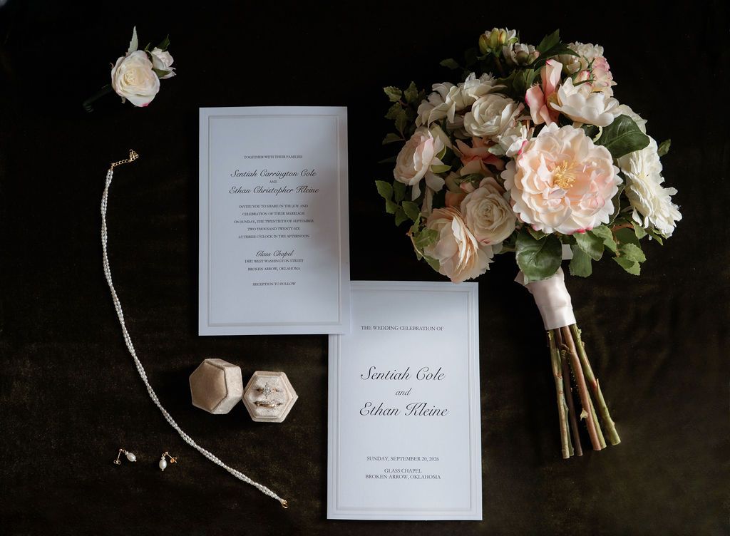 Elegant wedding flat lay with floral bouquet, invitation suite, ring box, and boutonniere on dark background.