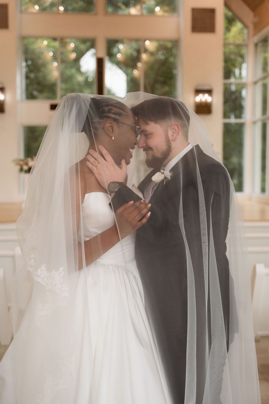 Interracial newlyweds sharing intimate moment under bridal veil in sunlit wedding chapel.