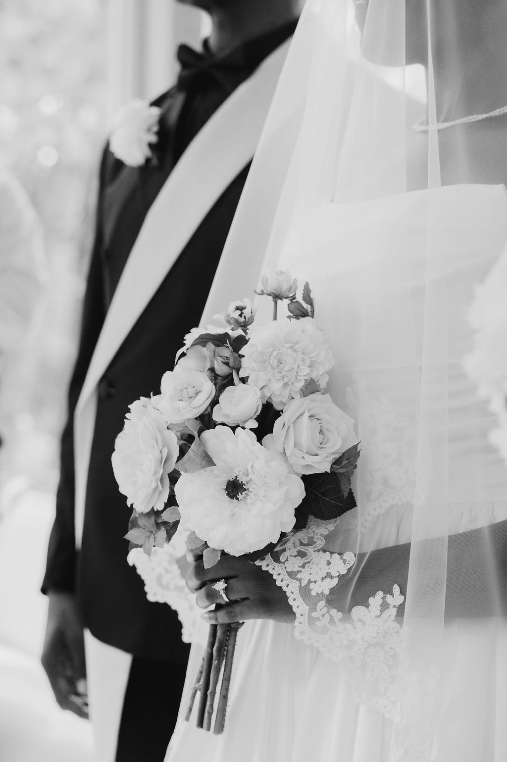 Black and white wedding photo of bride holding floral bouquet with roses beside groom in tuxedo.