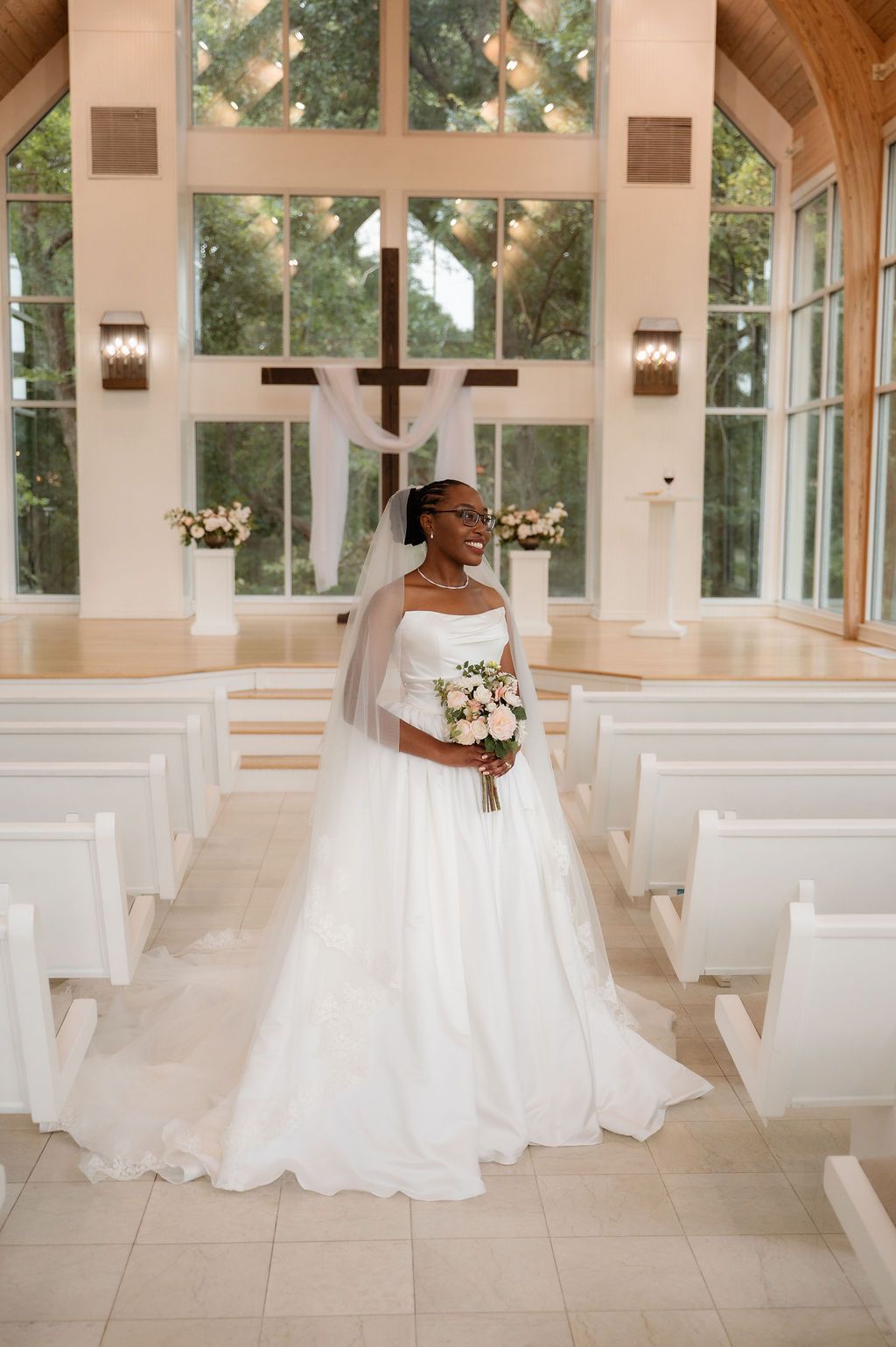 Bride in white ball gown holding bouquet stands in sunlit chapel aisle with cross and pews.