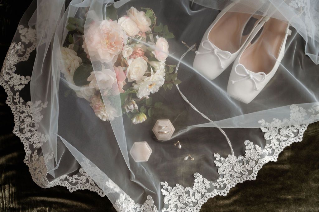 Bridal flat shoes, lace veil, pink flower bouquet, and rings arranged elegantly on a dark surface.