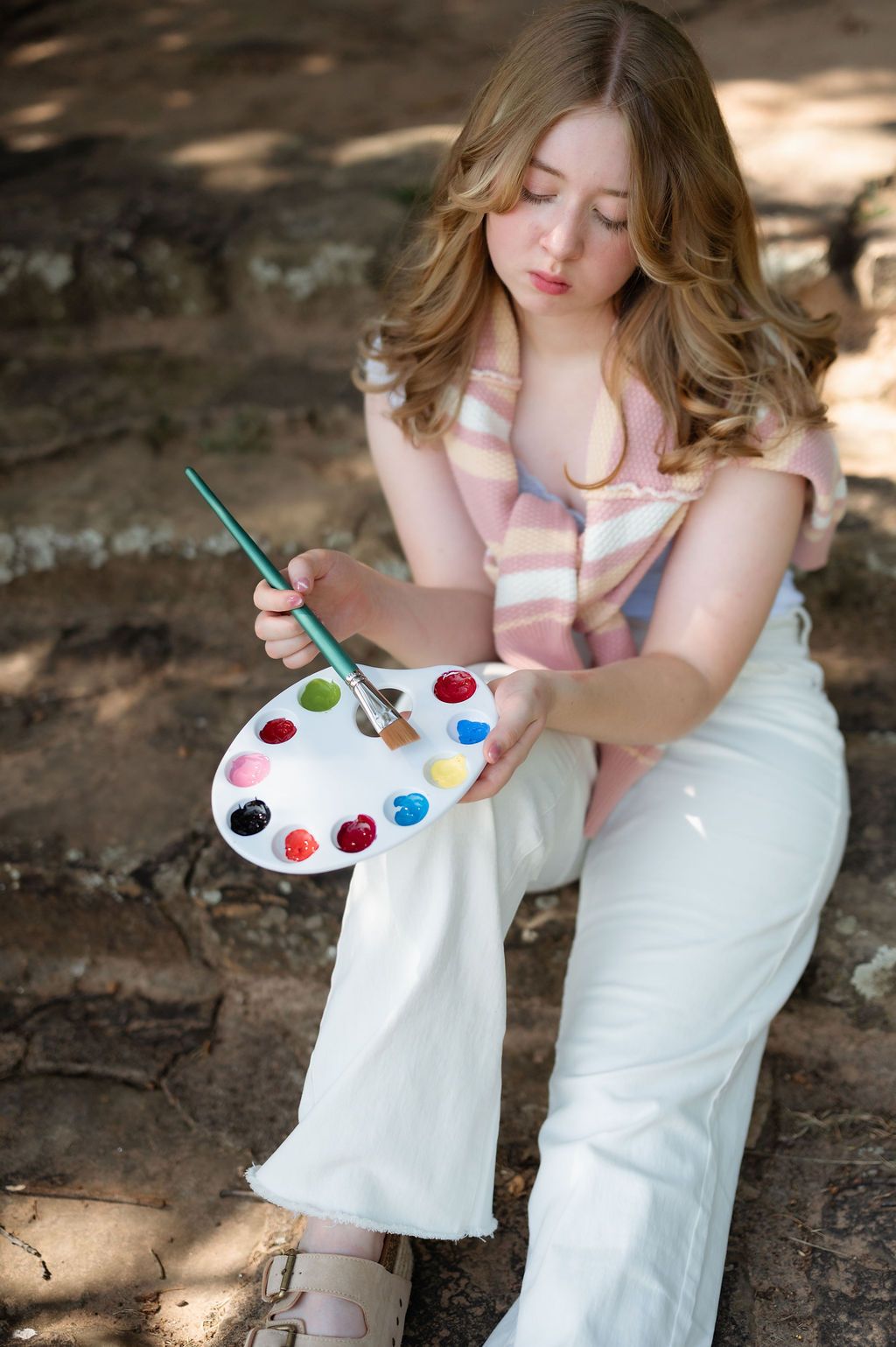 Senior girl with wavy hair holding a paint palette and brush while sitting on stone steps at Oklahoma Park