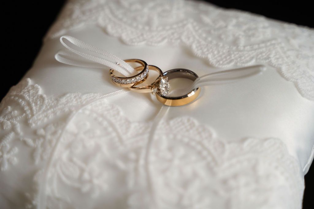 Two gold wedding rings with diamonds resting on a white lace ring bearer pillow.