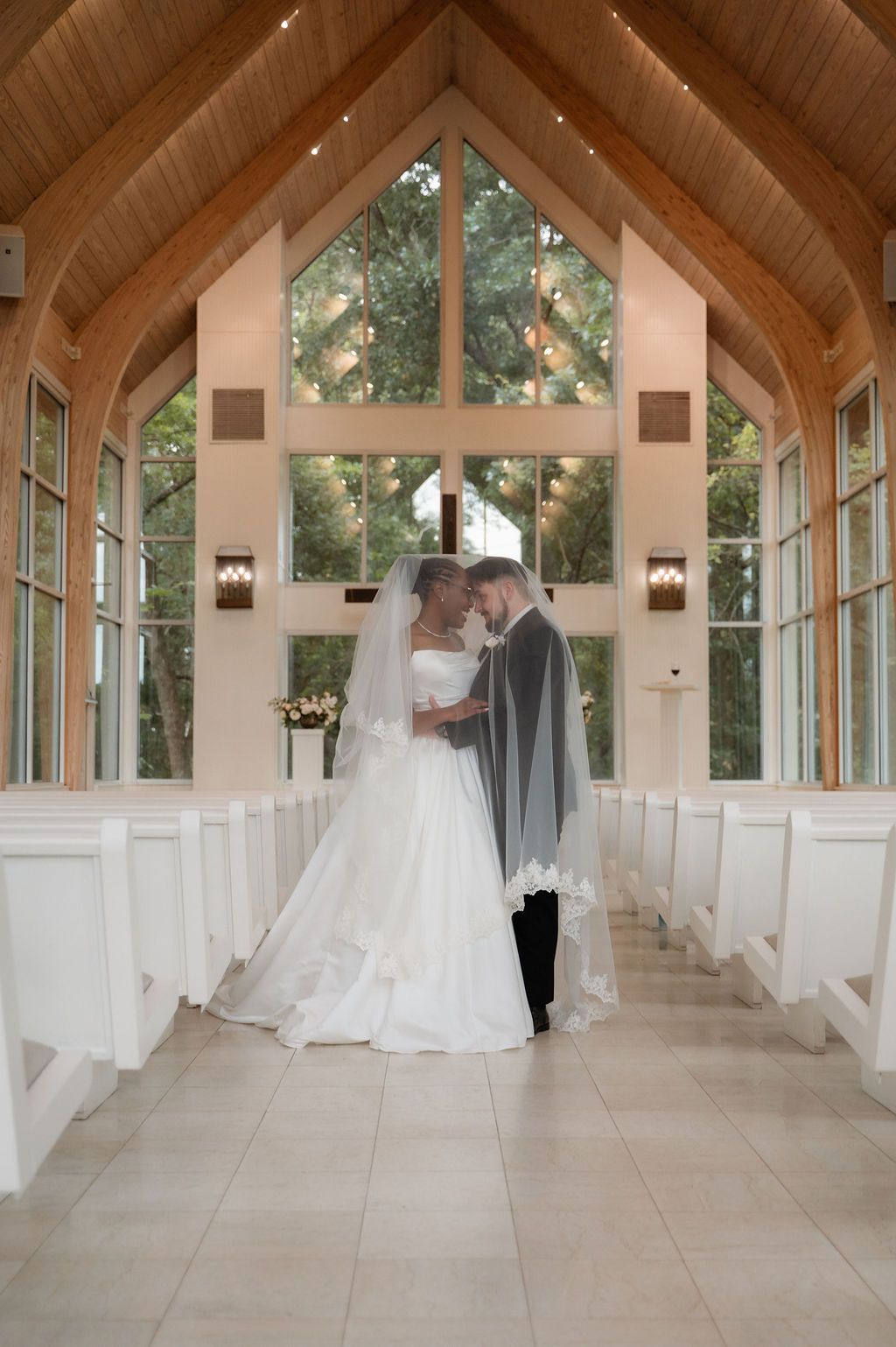 Bride and groom share intimate moment in elegant chapel with vaulted wood ceiling and white pews.