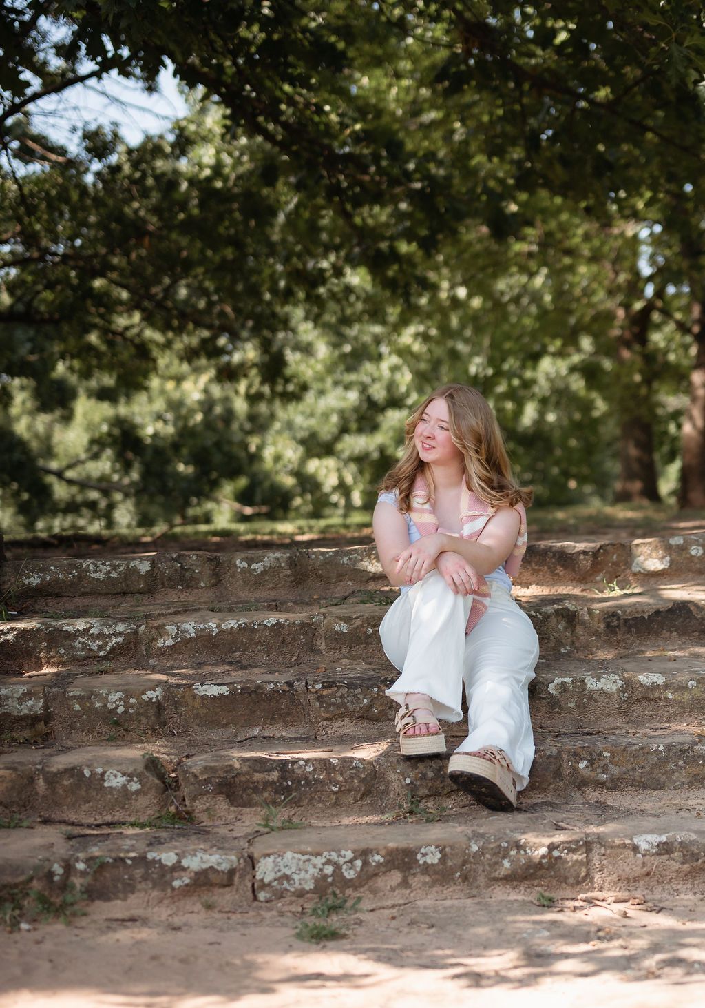 Smiling senior girl in white flared pants sitting on stone steps surrounded by lush green trees.