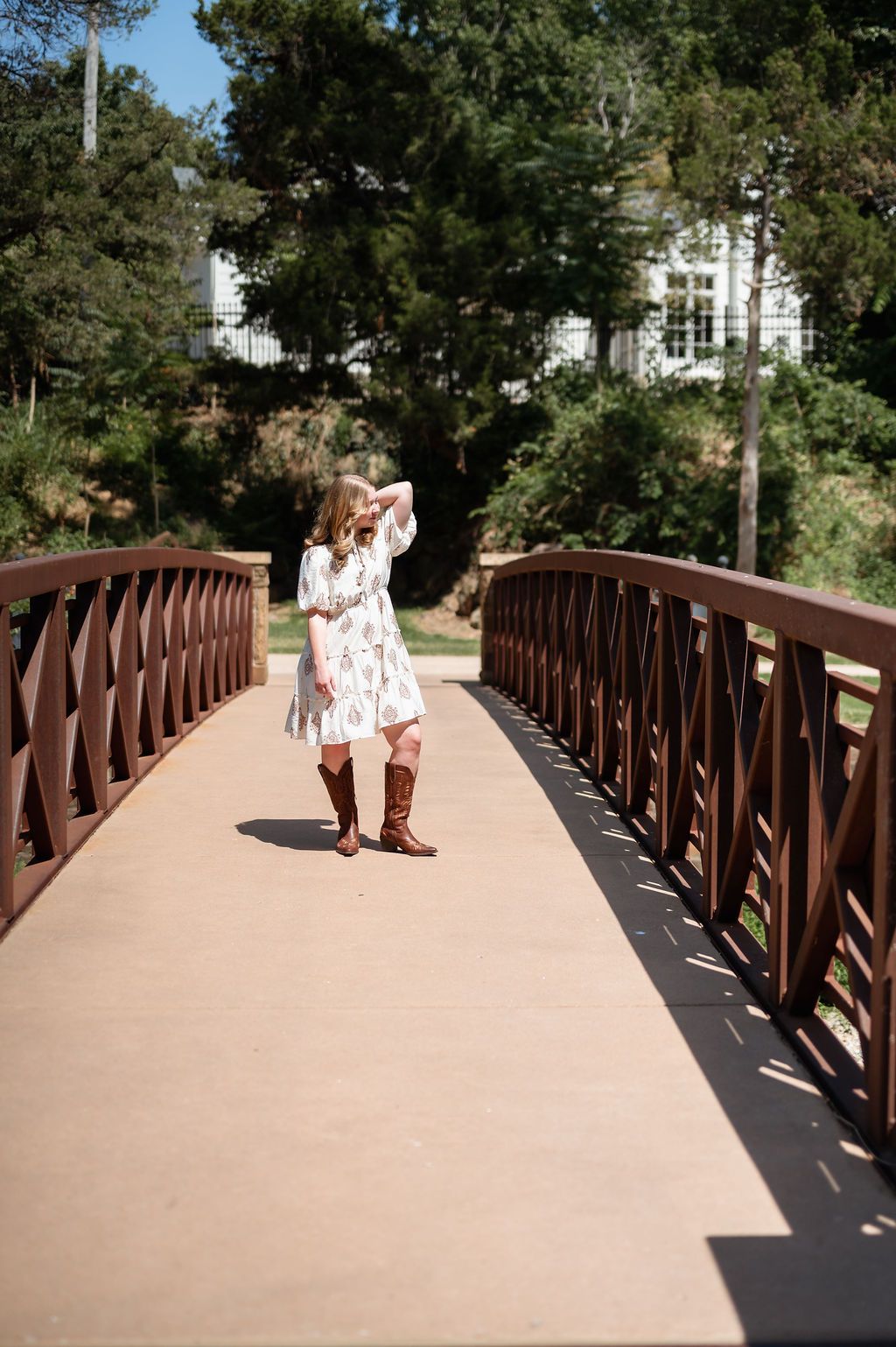 Young woman in white floral dress and brown boots poses on wooden bridge for senior portraits.