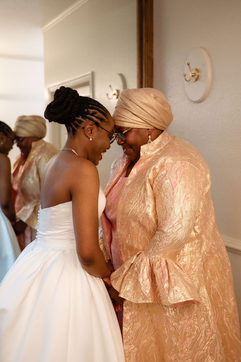 A bride in a white gown shares an intimate forehead touch with an older woman in a pink brocade outfit at a wedding.