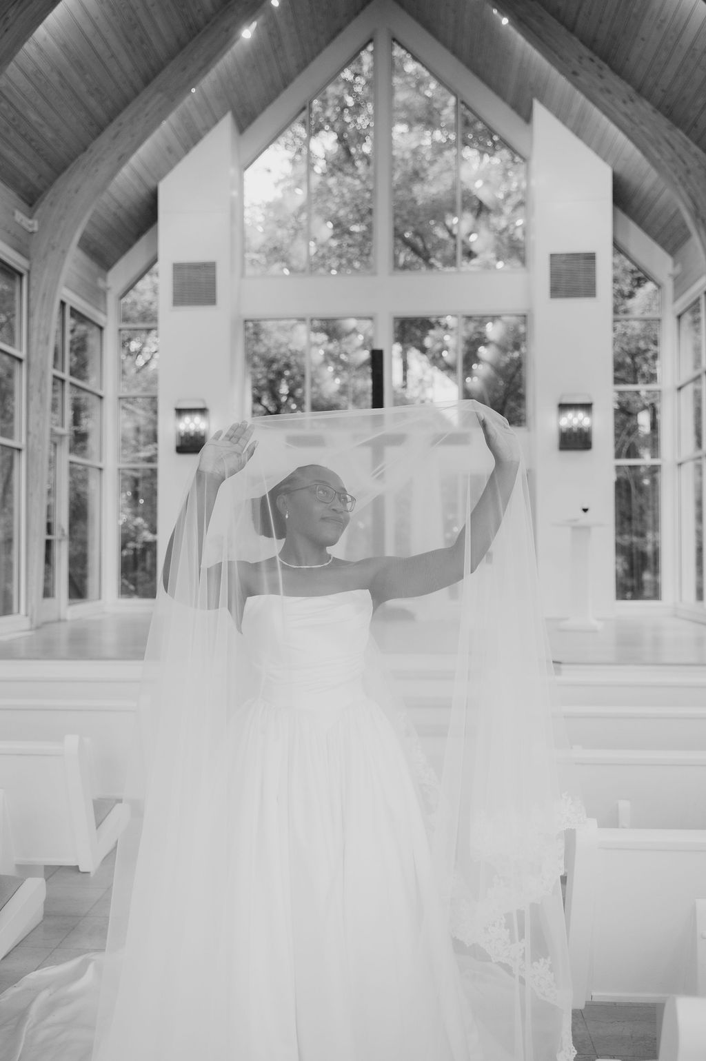 Black and white photo of a bride in a white gown lifting her veil inside a sunlit chapel with large windows.