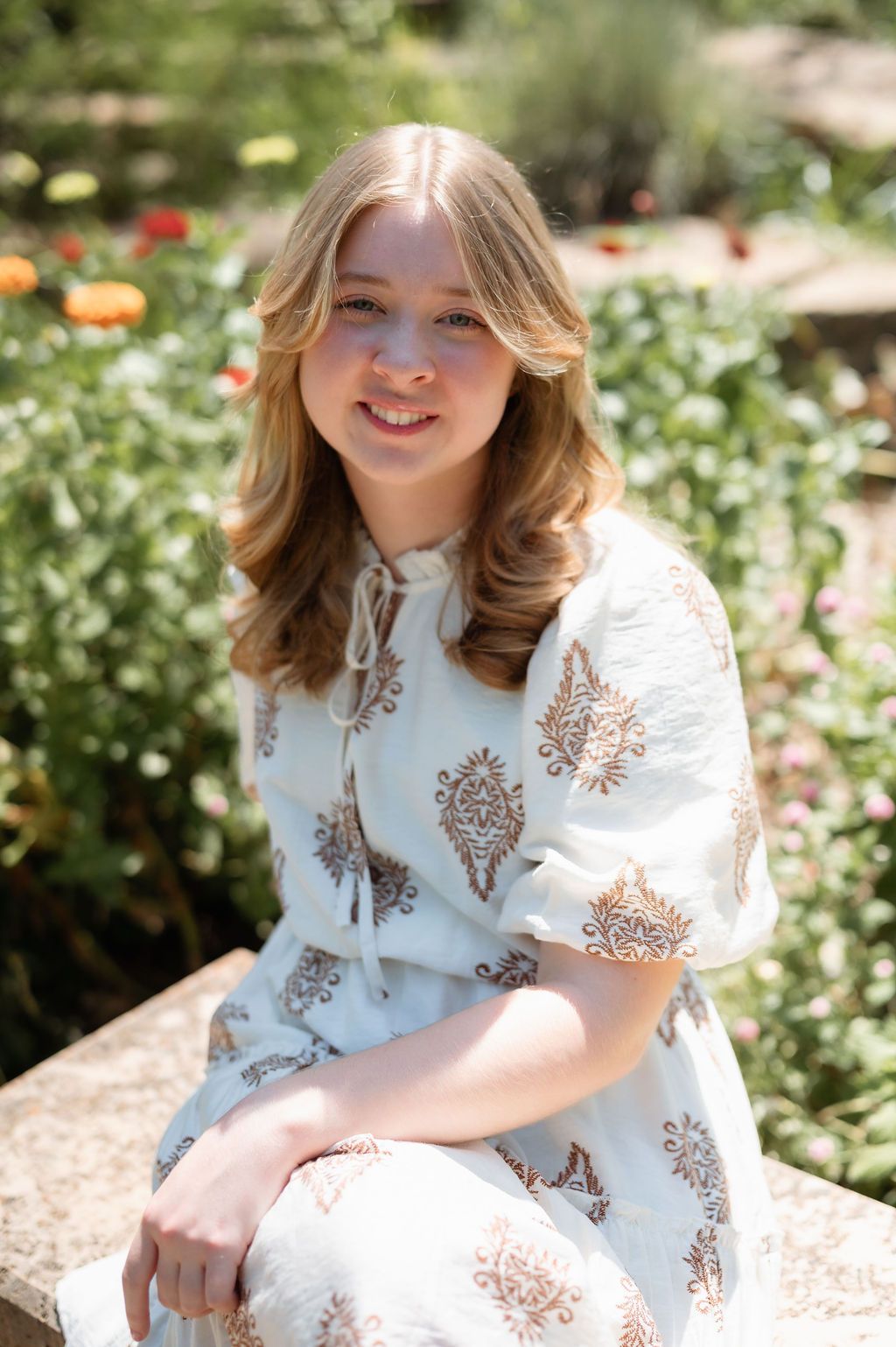 Smiling young woman in light blue paisley dress sits outdoors surrounded by colorful garden flowers.