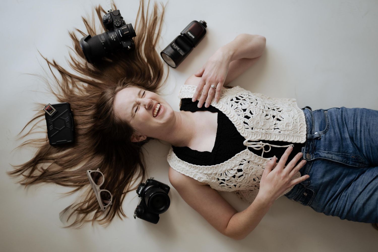 Young woman lying on floor laughing with cameras, lens, sunglasses, and phone surrounding her.