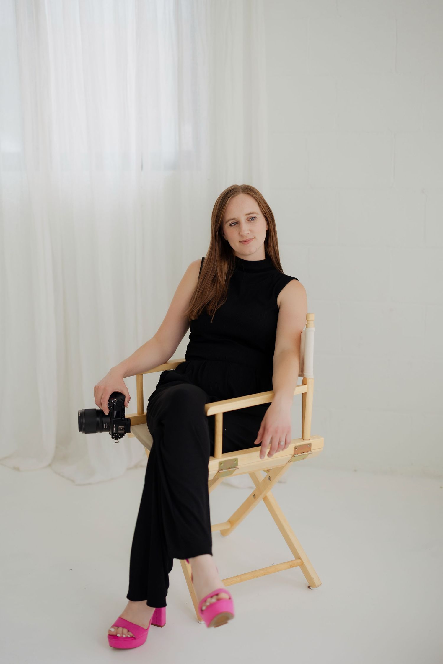 Woman in black outfit and pink sandals sits in a director's chair holding a camera in a white studio.
