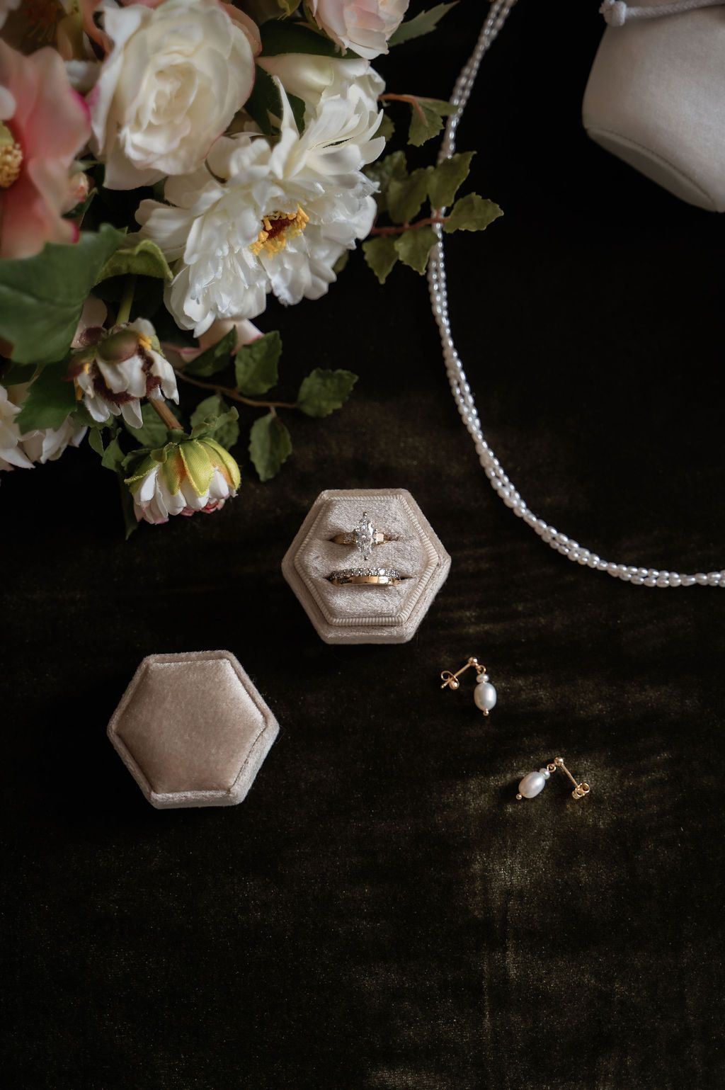 Elegant bridal flat lay with diamond rings in velvet box, pearl necklace, drop earrings, and pink floral bouquet on dark surface.
