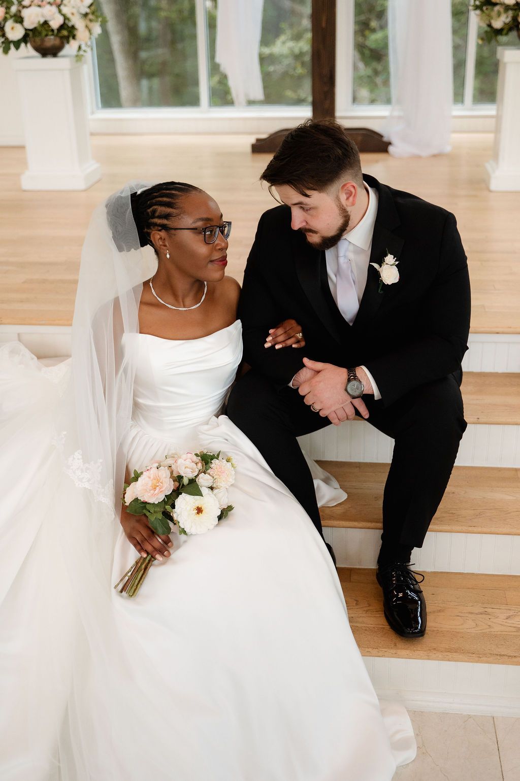 Bride in white gown and groom in black tuxedo sitting on chapel steps, gazing at each other.