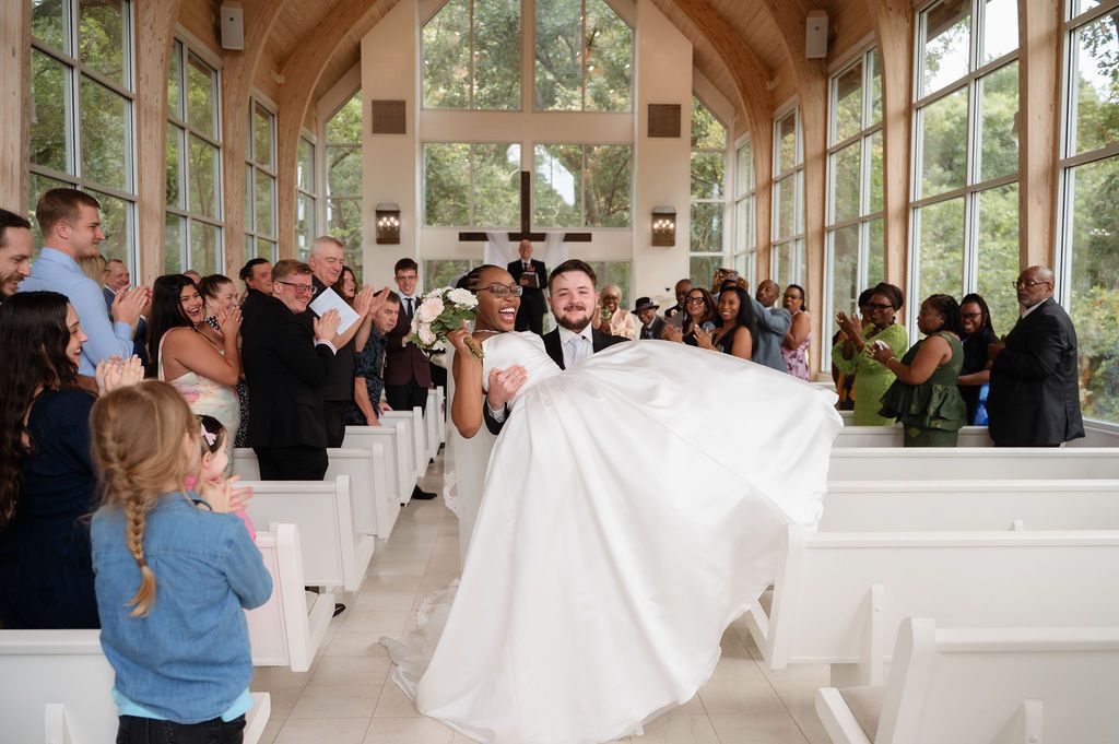 Joyful groom lifting bride in white ballgown as guests celebrate in sunlit chapel aisle.