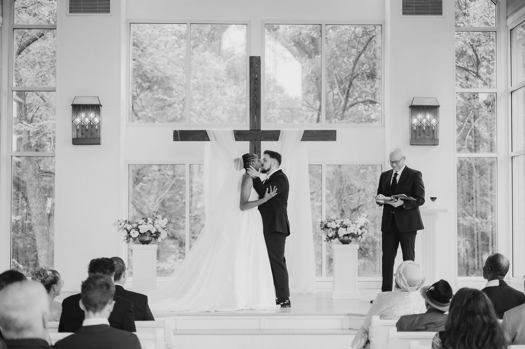 Black and white wedding ceremony photo of couple kissing at altar before cross in elegant chapel with guests seated.