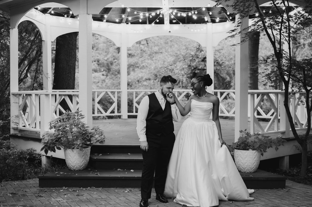 Black and white photo of a laughing bride and groom standing before a decorated gazebo at their outdoor wedding.