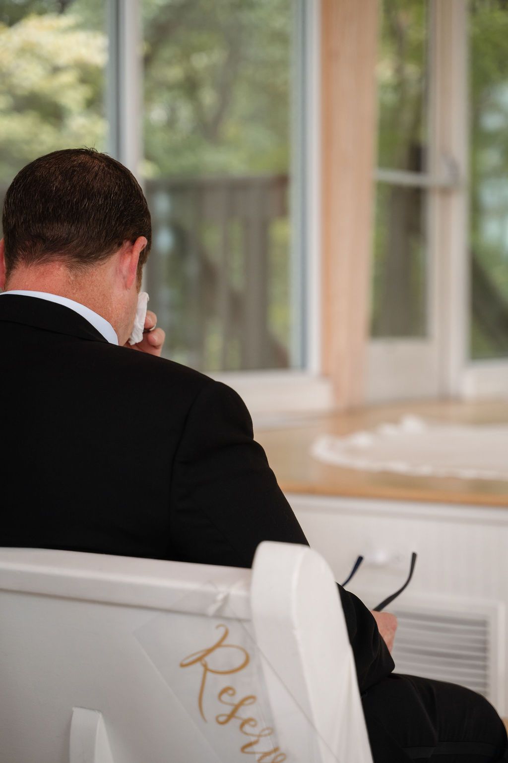 Emotional wedding guest in black suit wiping tears while seated in white reserved chair during ceremony.