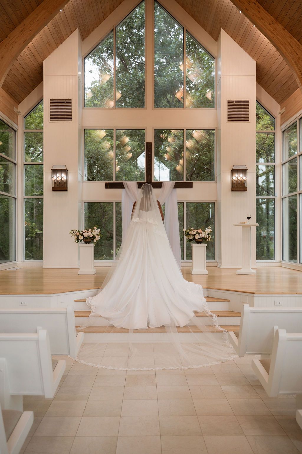 Bride in white gown and veil stands before wooden cross in sunlit chapel with large windows.