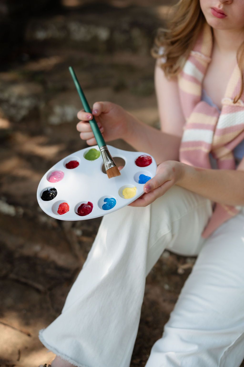 Close-up of hands holding a white paint palette with colorful paints and a green brush.