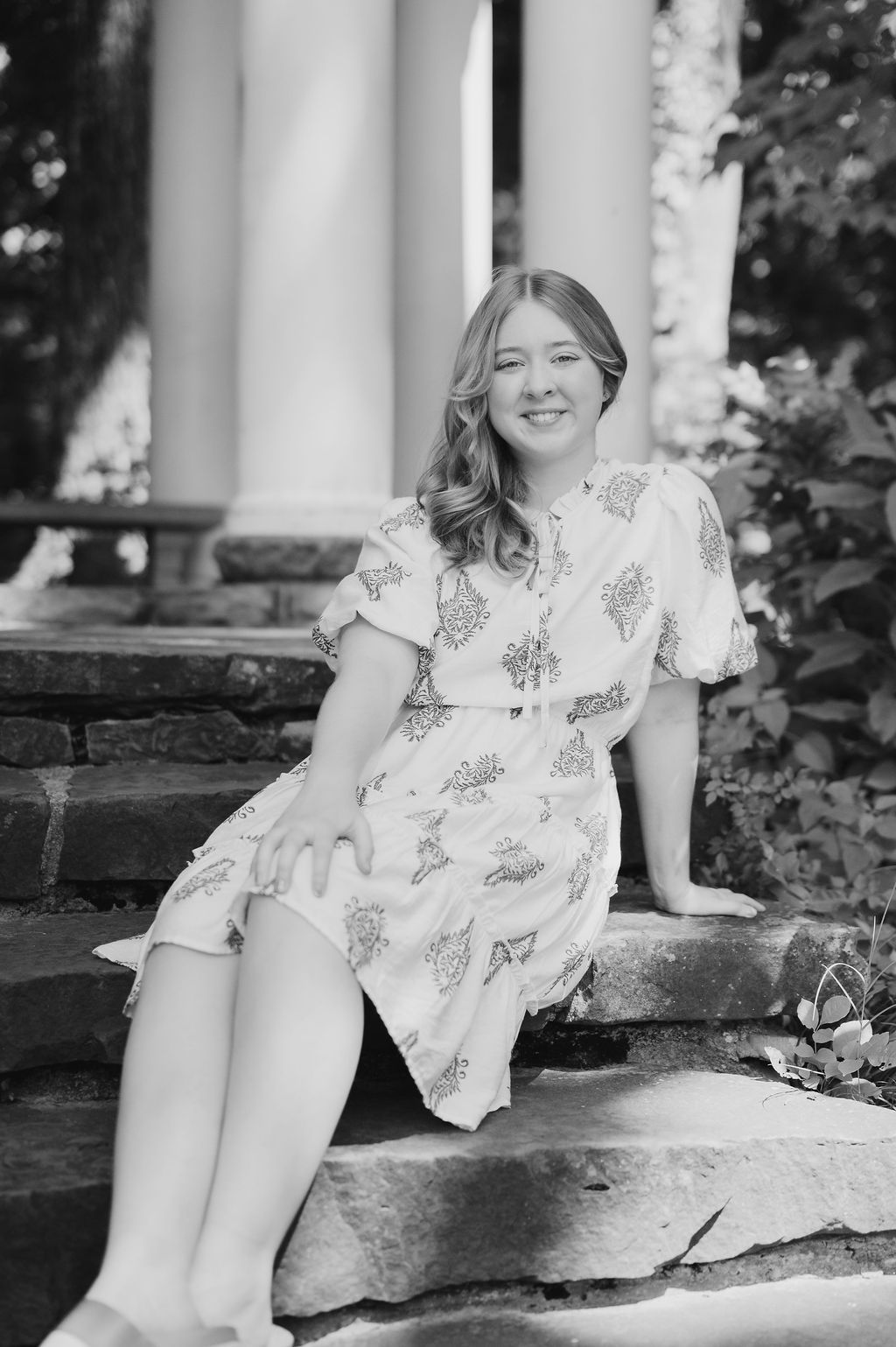 Black and white photo of laughing teen girl in patterned dress sitting on stone steps outdoors.