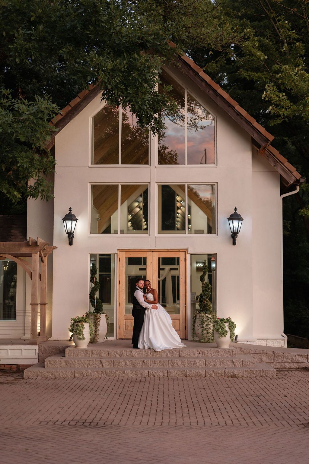 Bride and groom embracing at dusk before a white chapel with large wooden doors and warm glowing lanterns.