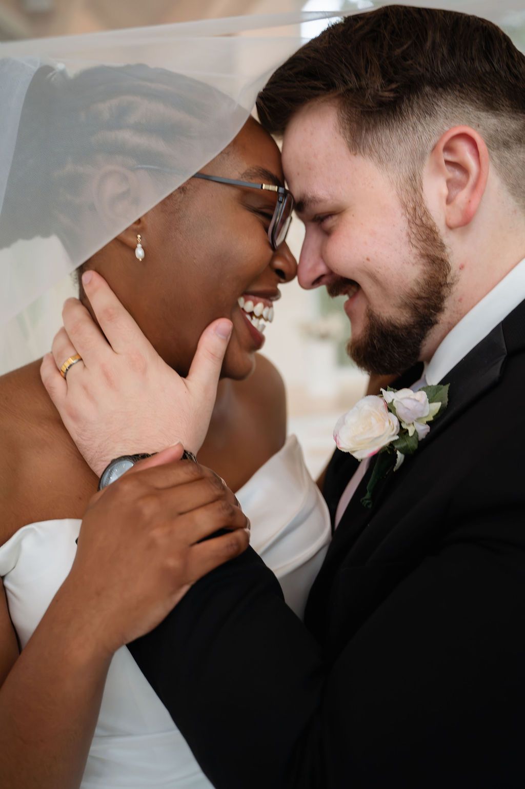 Joyful newlyweds touch foreheads under bridal veil, sharing a loving smile on wedding day.