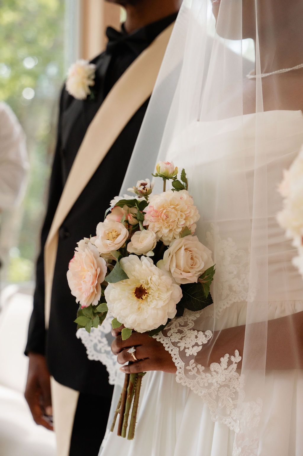 Bride holding elegant blush and white peony rose bouquet in lace wedding gown beside groom.